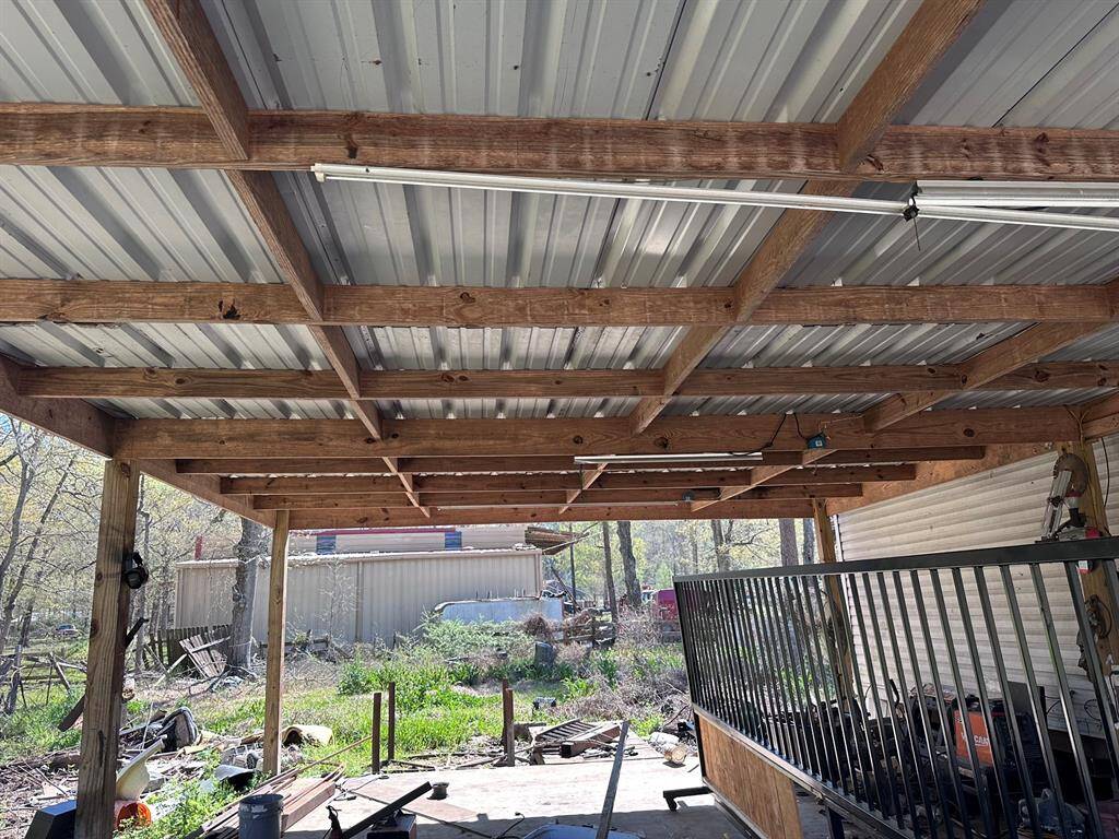 This photo shows a covered outdoor space with a metal roof and wooden beams. The area appears to be used for storage or work, with tools and equipment visible. The surroundings are grassy with some trees and a metal structure in the background.