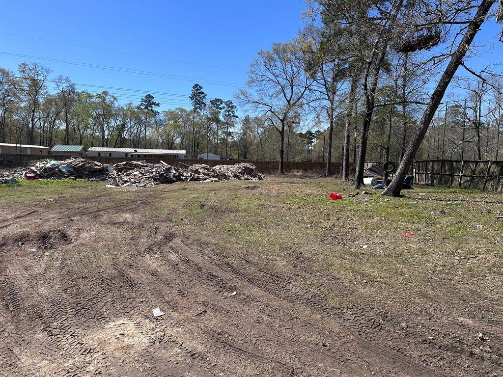 A spacious plot with scattered debris and some trees, bordered by a fence and neighboring buildings. Ideal for redevelopment with a natural backdrop.