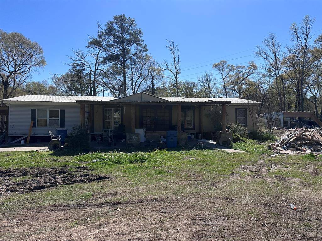 This property features a single-story home with a metal roof, set on a spacious lot with surrounding trees. The exterior shows potential for improvement and landscaping, with a covered front porch area. The yard has ample space but requires some cleanup and maintenance.