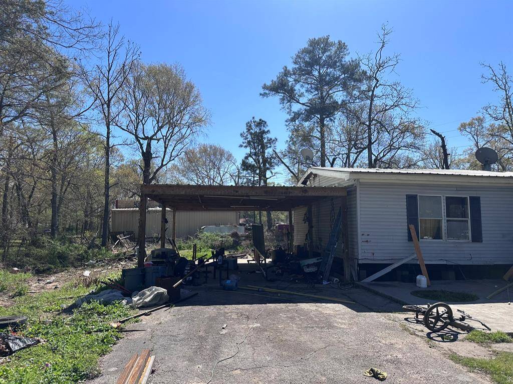 This photo shows a single-story home with a carport, set in a wooded area. The property appears to have some clutter and could use maintenance, but offers ample outdoor space with potential for improvement.