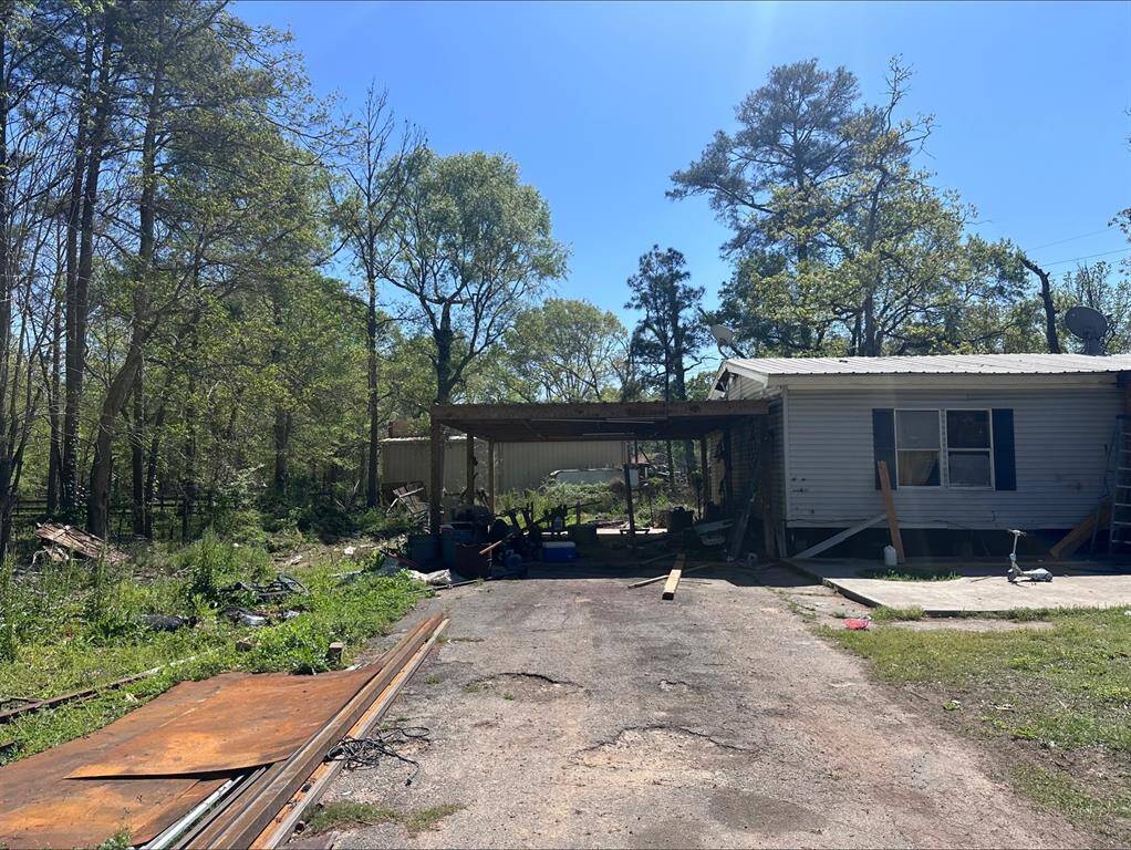 This property features a rustic one-story home with an attached carport, set amidst a wooded area. The exterior shows signs of wear and there is some debris present, offering a potential fixer-upper opportunity.
