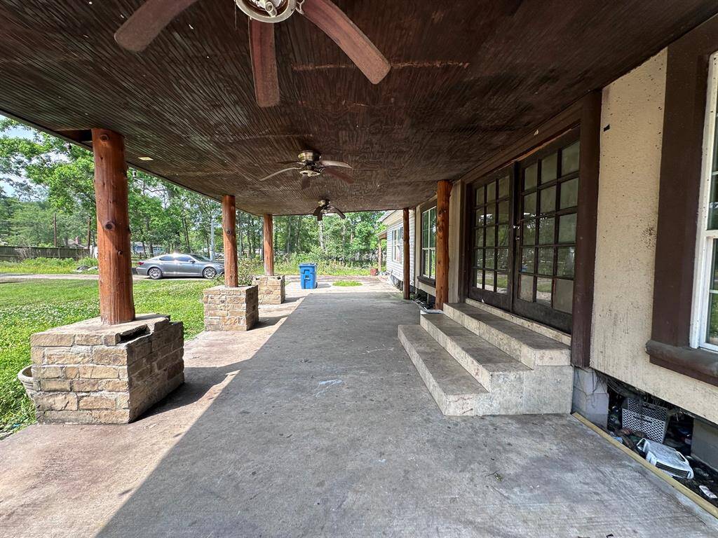 Spacious covered patio with rustic wooden beams and ceiling fans, featuring stone pillars and steps leading to large glass doors. Ideal for outdoor relaxation and entertaining.