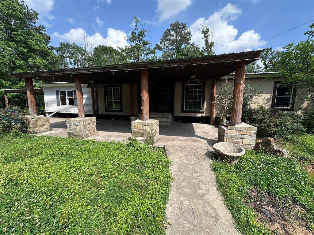 A rustic home with a covered front porch featuring wooden beams and stone pillars. The lush greenery and overgrown grass add a natural charm to the property.