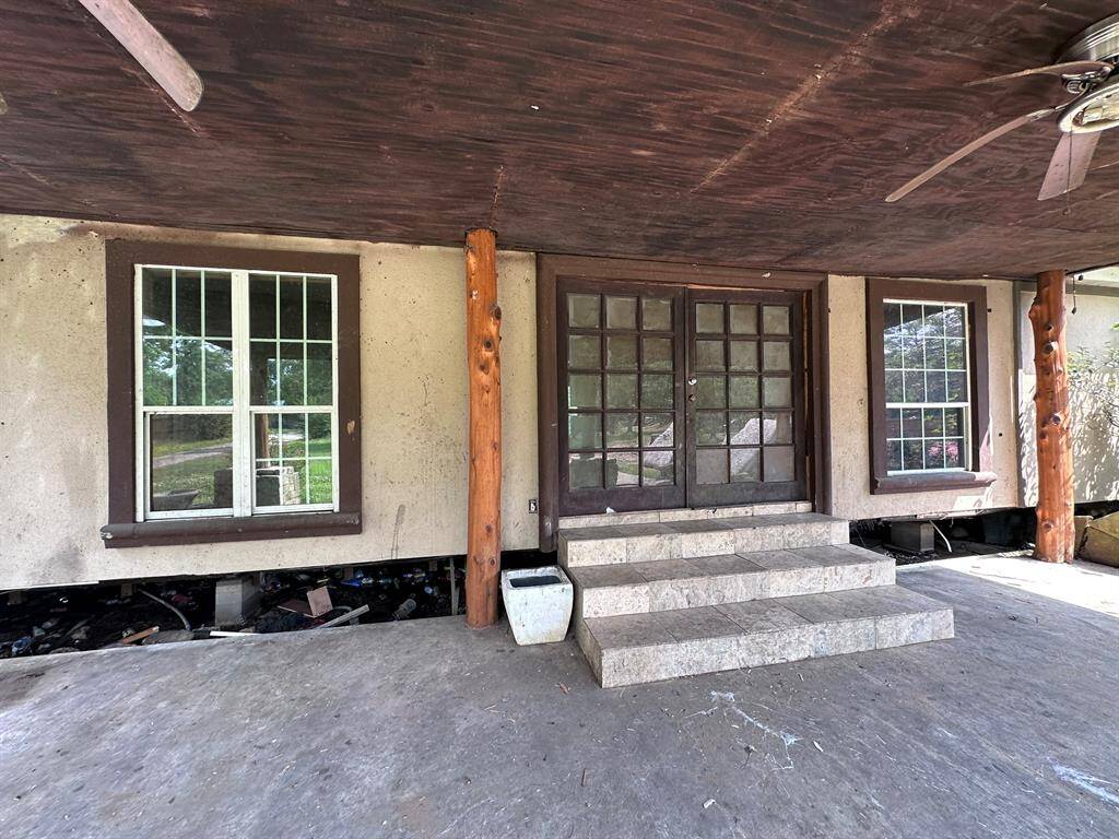This photo shows a rustic front porch with a wooden ceiling, stone steps leading to double doors, and two large windows. There are wooden support beams and a ceiling fan, creating a cozy, shaded entryway.