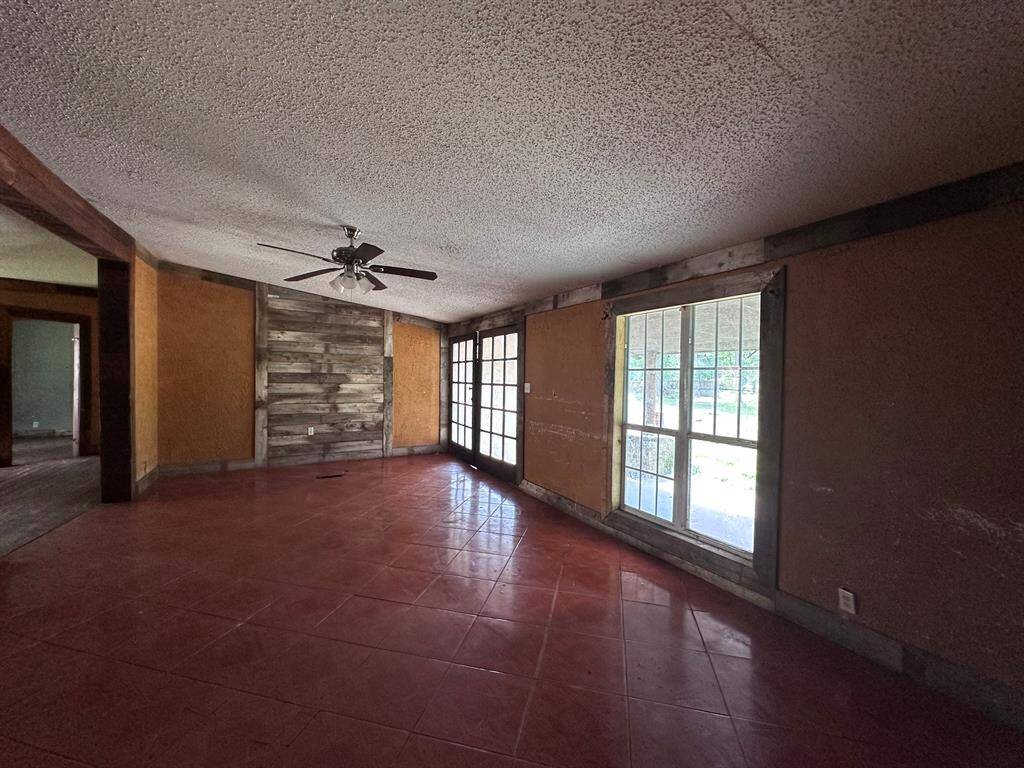 This spacious room features large windows for ample natural light, a ceiling fan, and rustic wood accents on one wall. The red tile flooring adds warmth and character to the space, making it ideal for a cozy living area.