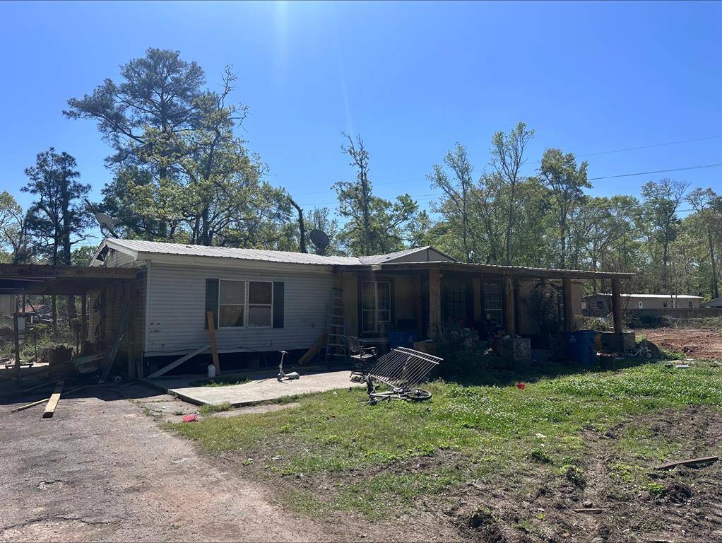 This photo shows a single-story home with a metal roof and a covered porch. It's situated on a spacious lot with surrounding trees, though the yard appears a bit cluttered. The exterior looks rustic, with potential for improvement and personalization.