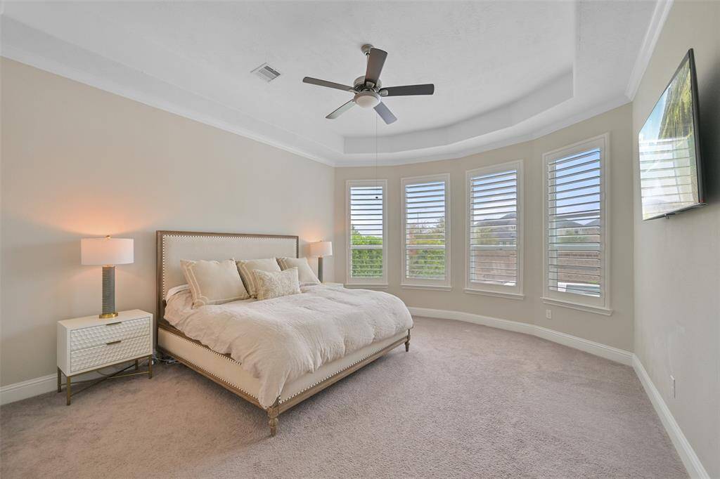 The spacious primary suite offers a serene retreat with a wall of windows, enhanced by added plantation shutters for both privacy and natural light. The room features plush low-profile carpet, a tray ceiling that adds architectural detail, and a ceiling fan/light fixture for comfort and style.