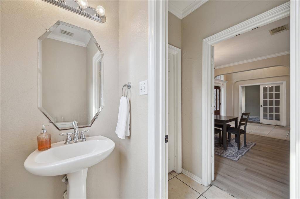 This photo shows a well-litpowder bathroom with a pedestal sink and octagonal mirror. Adjacent is a dining area with a table and chairs, featuring wood and tile flooring and French doors.