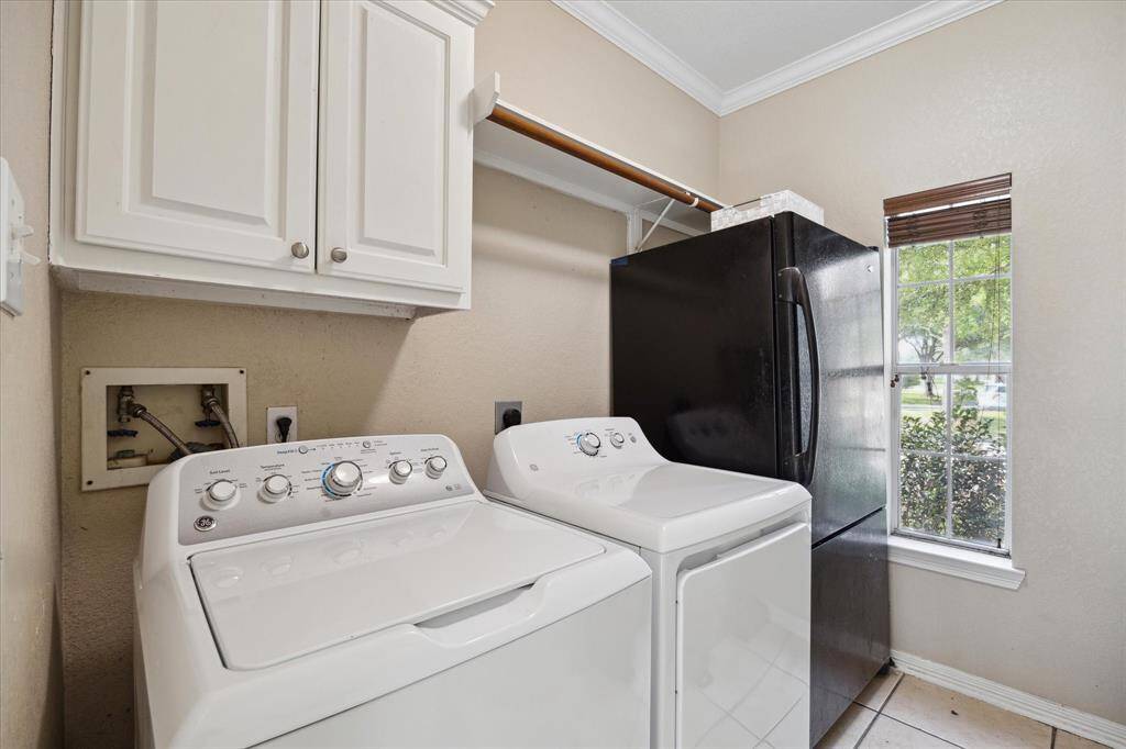 This laundry room features a washer and dryer, upper storage cabinets, a large black refrigerator, and a window with natural light.