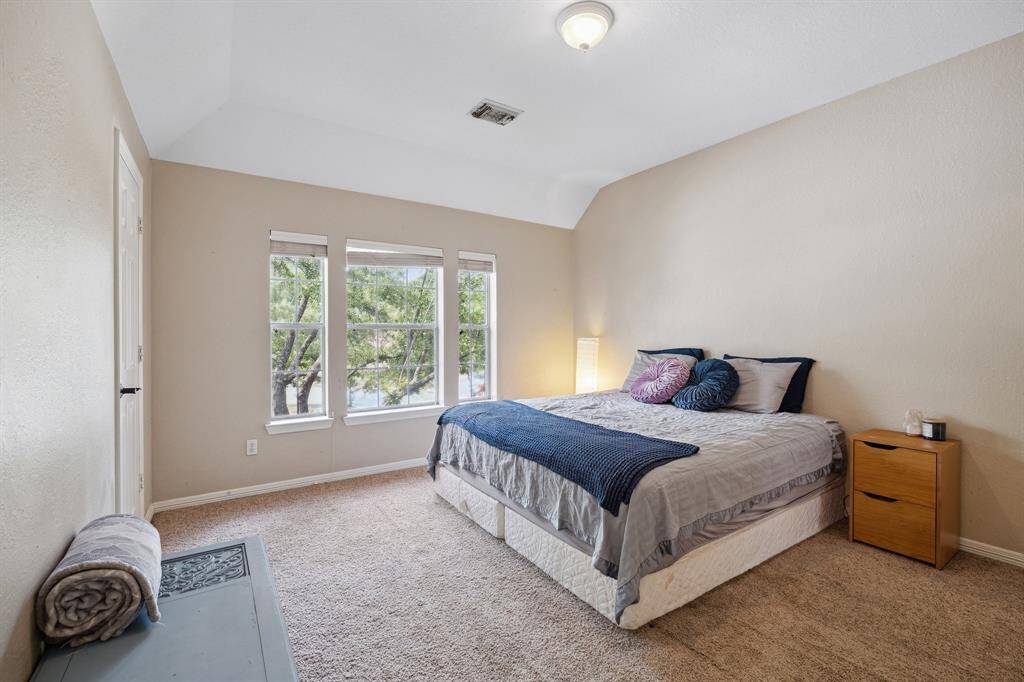Bright bedroom with neutral walls, featuring a large bed, bedside table, and windows offering natural light and views of greenery. Plush carpeting adds warmth and comfort. This is a large secondary bedroom off the jack and jill bathroom.