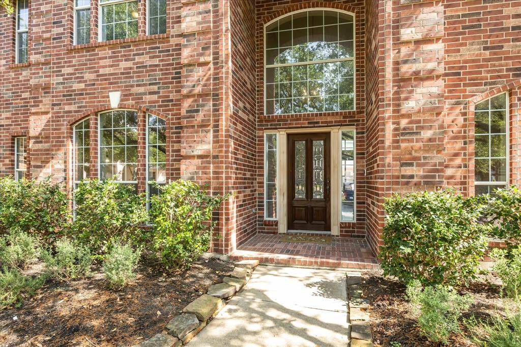 This photo showcases the entrance of a brick home with large, arched windows and a dark wooden front door. The pathway is lined with well-maintained shrubs and plants, leading to the inviting entryway. Roof 9 years old. AC recently serviced and tuned up last week.