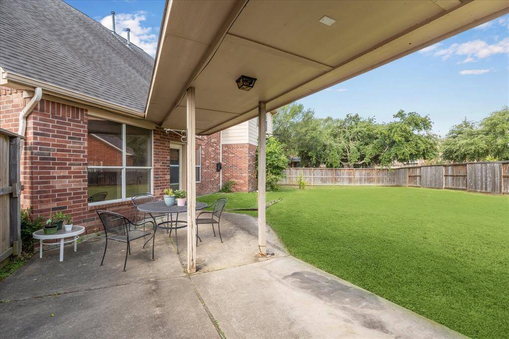 This photo showcases a backyard with a covered patio/breezeway and a seating area, set against a well-maintained lawn. The breezeway leads to a double garage. The space is bordered by a wooden fence and features a brick exterior, providing a private and inviting outdoor area.