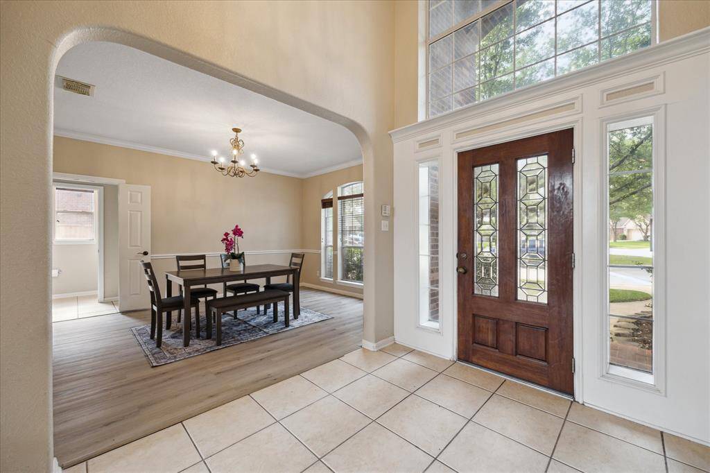 This inviting entryway features a wooden front door with decorative glass panels, high ceilings, and ample natural light. It opens into a spacious dining area with elegant flooring and a classic chandelier, creating a warm and welcoming atmosphere.