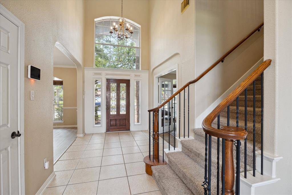 This inviting foyer features high ceilings and a grand staircase with wooden railings. A large window above the front door allows natural light to fill the space, showcasing the elegant chandelier and tiled flooring. An open archway leads to adjacent living areas, enhancing the home's spacious feel.