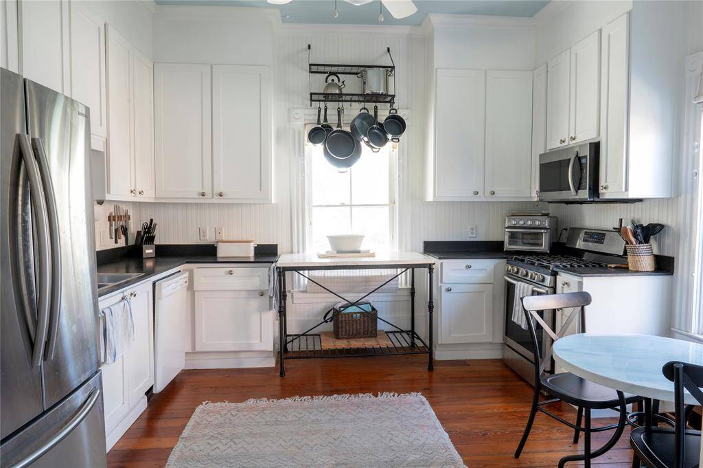 This open kitchen features white cabinetry, stainless steel appliances, and a cozy dining area. It includes a hanging pot rack, wooden floors, and ample natural light, creating a bright and welcoming space.