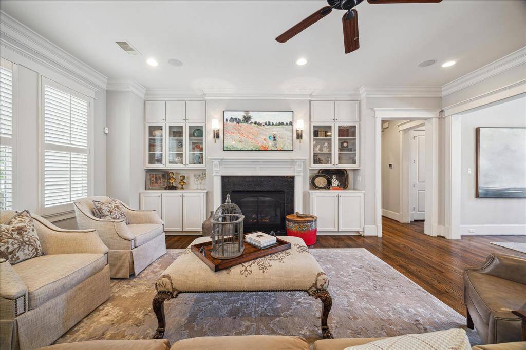 Inviting living room centered around a gas fireplace with detailed wood mantel and granite surround. Flanked by custom built-ins with glass-front cabinetry and accent lighting, the space blends function and design. Crown molding, oak floors, and natural light from plantation-shuttered windows complete this warm and elegant gathering area.