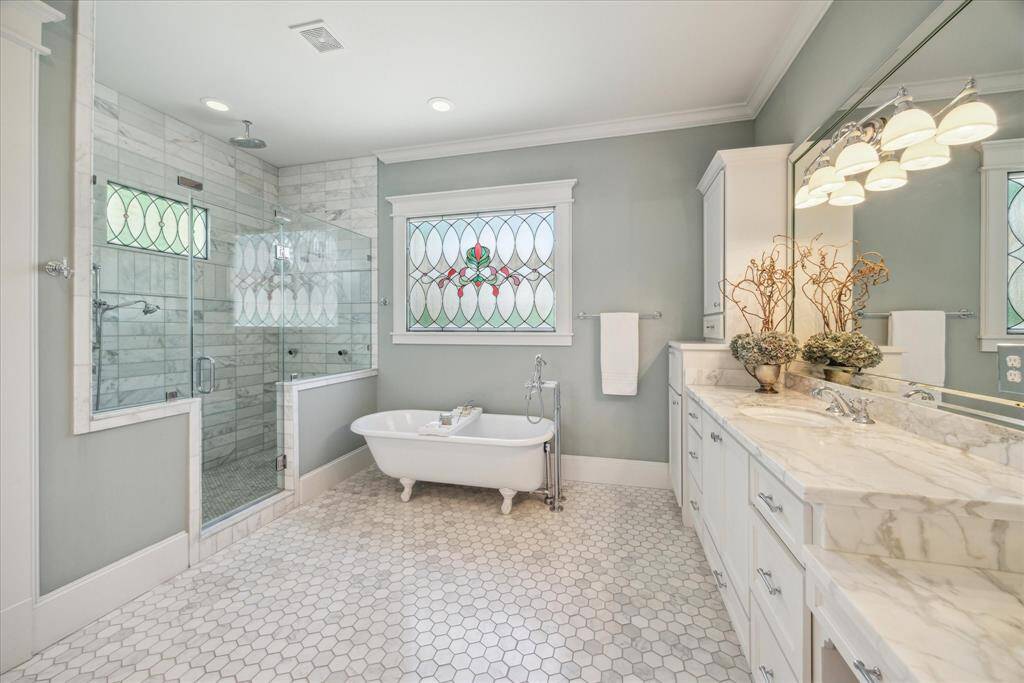The primary bath features a dual-head marble-clad shower, expansive double vanity, and the original circa 1910s± clawfoot tub. Soft natural light filters in, highlighting the elegant hexagonal marble floors and creamy veined countertops. Thoughtful built-ins and polished chrome fixtures complete the look.