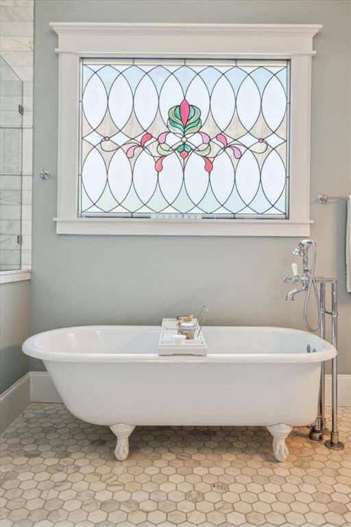 Framed beneath an artisan-crafted stained glass window—featuring rose, jade, and amber tones set in a vintage oval lattice—the original 1910s± clawfoot tub anchors the space with historic elegance. Its cast iron form and sculpted feet speak to a bygone era of craftsmanship, while the Rohl Viaggio polished chrome tub filler and tray invite moments of pure, indulgent calm.
