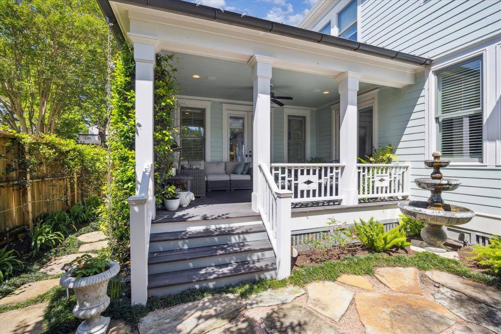 Framed by crisp white columns and intricate railing details, the covered side porch is a seamless extension of the home’s Southern charm. A soft blue beadboard ceiling adds vintage authenticity, while climbing jasmine and lush container plantings bring life and color to the space.