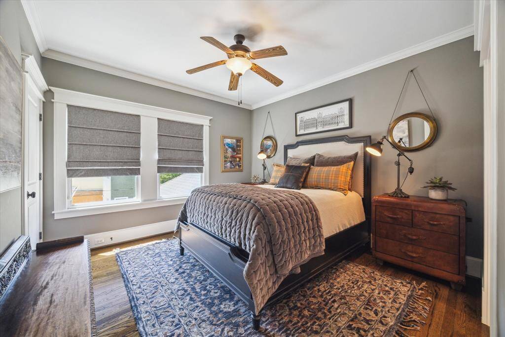 Tailored, textured, and perfectly balanced—this en-suite secondary bedroom proves that design is in the details. Large windows flood the space with natural light while contrasting beautifully against the warm, slate-toned walls. Roman shades add sophistication and softness, while wide crown molding and rich hardwoods deliver a grounded, architectural feel. A private bath is attached.
