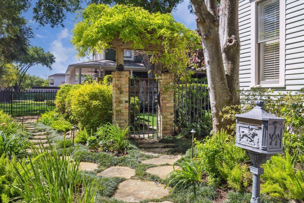 A winding flagstone path leads to a charming brick-and-iron gate, softly framed by mature greenery and climbing vines. The arbor overhead is draped in wisteria, creating a natural canopy and quiet transition from the front walk to the private garden beyond. The surrounding landscape, thoughtfully designed by Moss Landscaping, blends native plants and ornamental grasses for year-round interest. A vintage cast-iron mailbox adds a touch of historic character, tying together the home’s architectural past.
