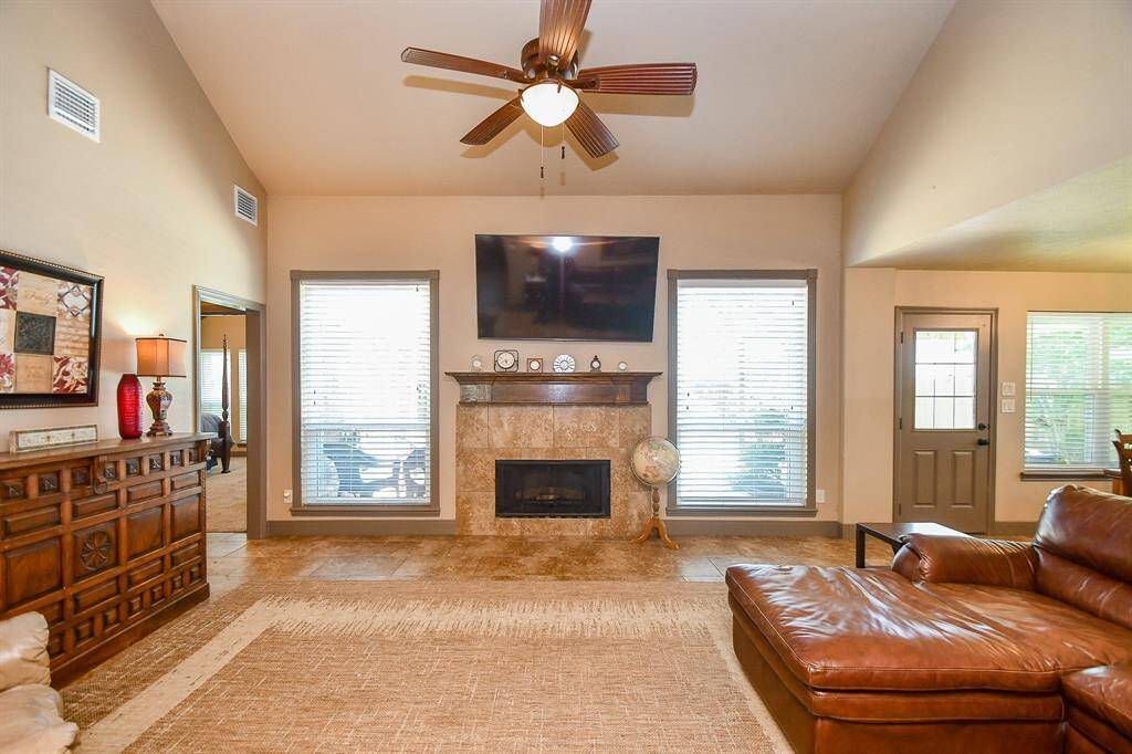 This spacious living room features a high ceiling with a ceiling fan, a cozy fireplace with a stone surround, and large windows that provide ample natural light. The room offerrs a greta view to the backyard paradise creating a warm and inviting atmosphere.
