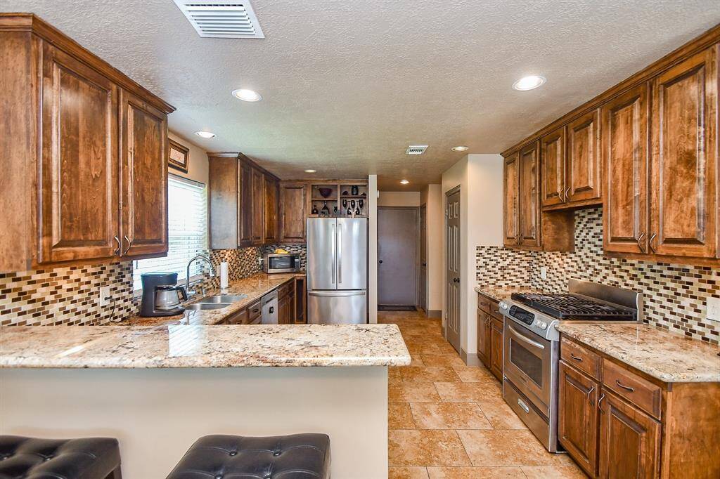 This kitchen features warm wooden cabinetry, granite countertops, and a tiled backsplash. It includes stainless steel appliances, a gas stove, and ample storage space. The open layout and recessed lighting create a bright, inviting atmosphere.