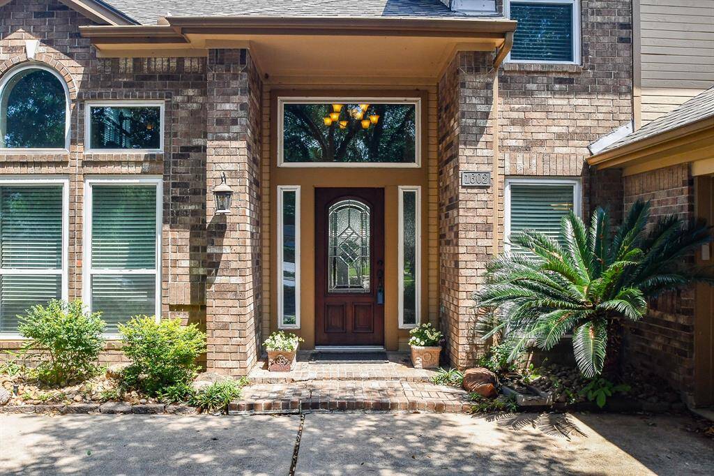 Charming brick home exterior with a welcoming front porch, elegant windows, and lush landscaping. The entryway features a stylish wooden door with decorative glass, framed by tall sidelights.