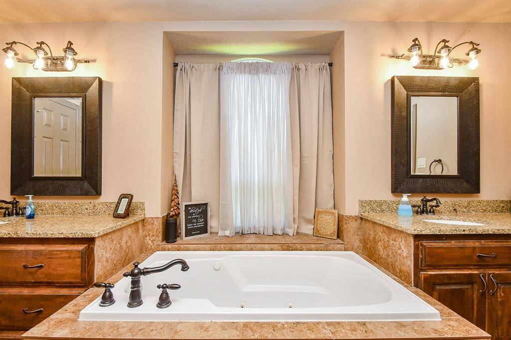 A luxurious bathroom featuring a large soaking/jetted tub set in a stone-tiled surround, flanked by dual vanities with granite countertops and framed mirrors. Warm lighting and rich wood cabinetry enhance the elegant ambiance.