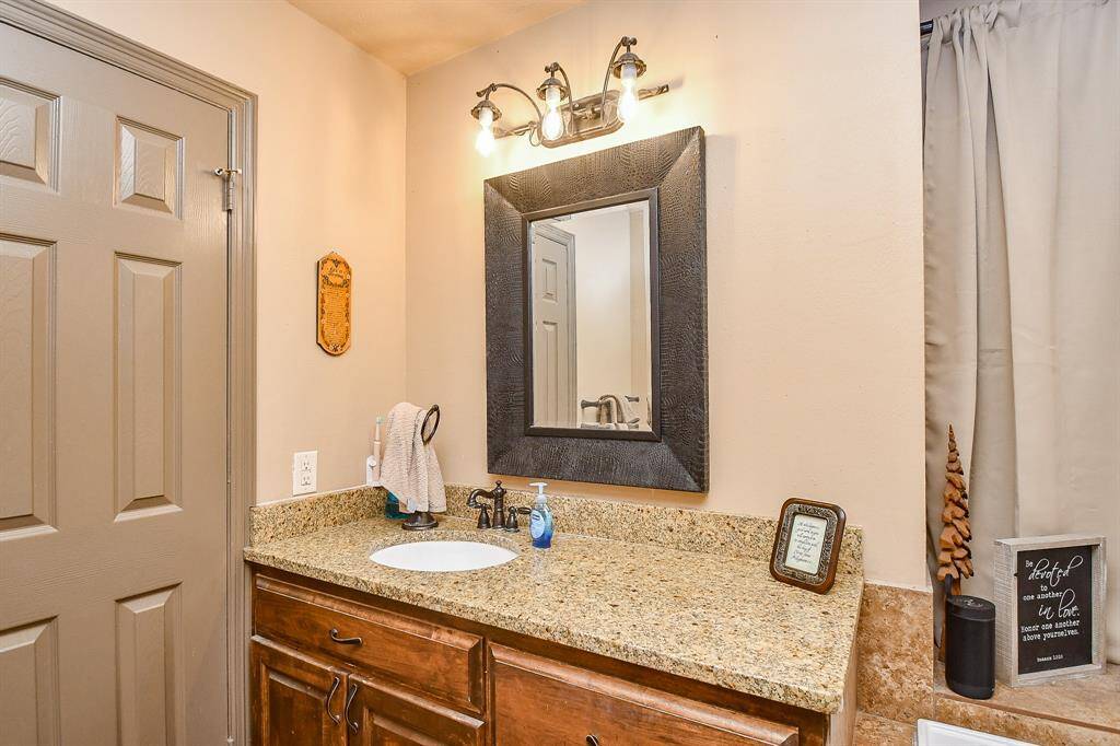 This area of the primary bathroom features a granite countertop with a single sink, wooden cabinetry, and a large framed mirror with overhead lighting. The neutral tones and decorative accents create a warm, inviting atmosphere.