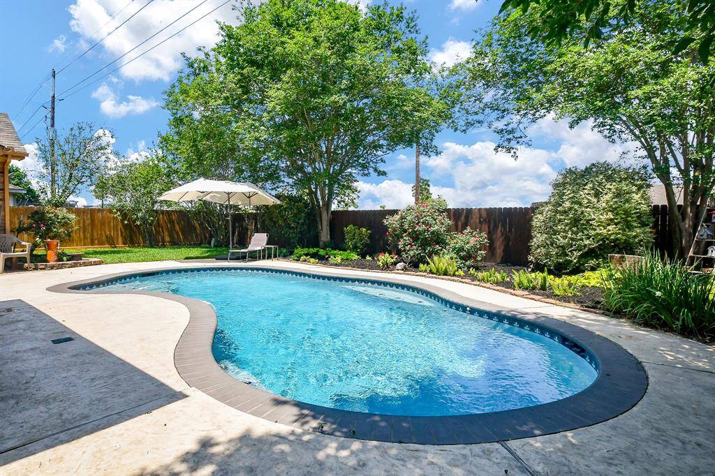 This backyard features a beautifully landscaped area with a freeform pool, surrounded by lush greenery and a wooden fence for privacy. A patio area large enough for lounge chairs and large umbrella to offer a perfect spot for relaxation and outdoor enjoyment.