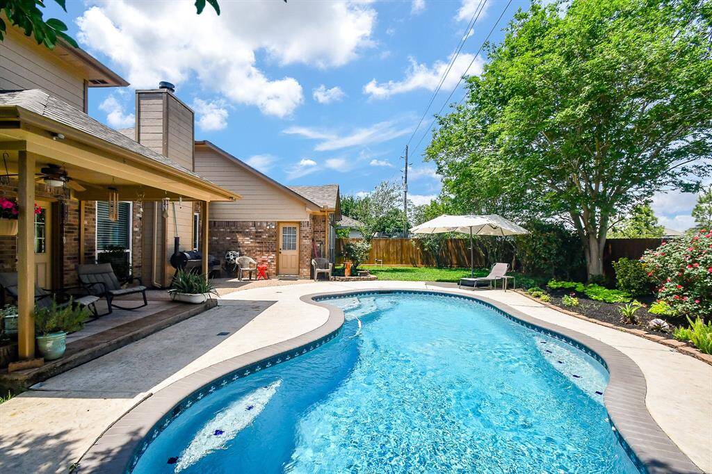 This photo showcases a spacious backyard featuring a curvy, inviting pool surrounded by a well-maintained patio and lush landscaping. A covered patio offers shade and relaxation, perfect for entertaining or enjoying sunny days.