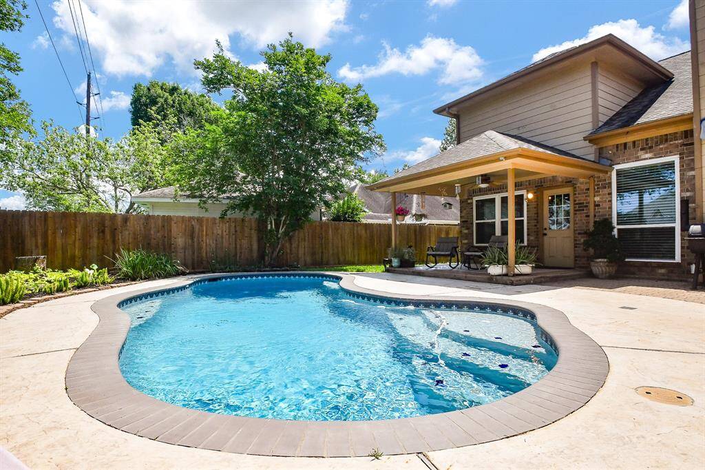 This photo showcases a charming backyard with a uniquely shaped in-ground pool, surrounded by a spacious patio area. The home features a cozy covered patio, perfect for outdoor dining or lounging, and is bordered by a privacy fence and lush greenery, creating a serene and inviting outdoor space.