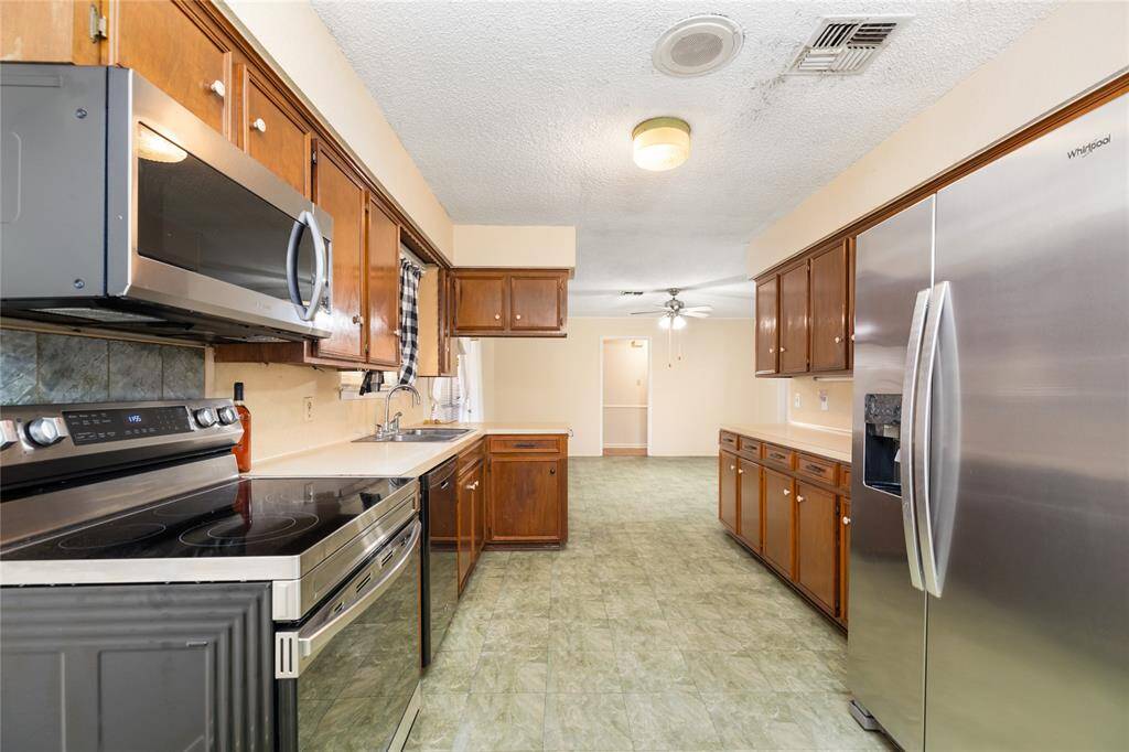 The kitchen boasts stainless steel appliances, rich wooden cabinets, and a spacious layout with plenty of counter space for cooking and entertaining. A ceiling fan enhances the bright, airy feel, while the open design seamlessly connects to a sunlit area, creating a warm and inviting atmosphere.