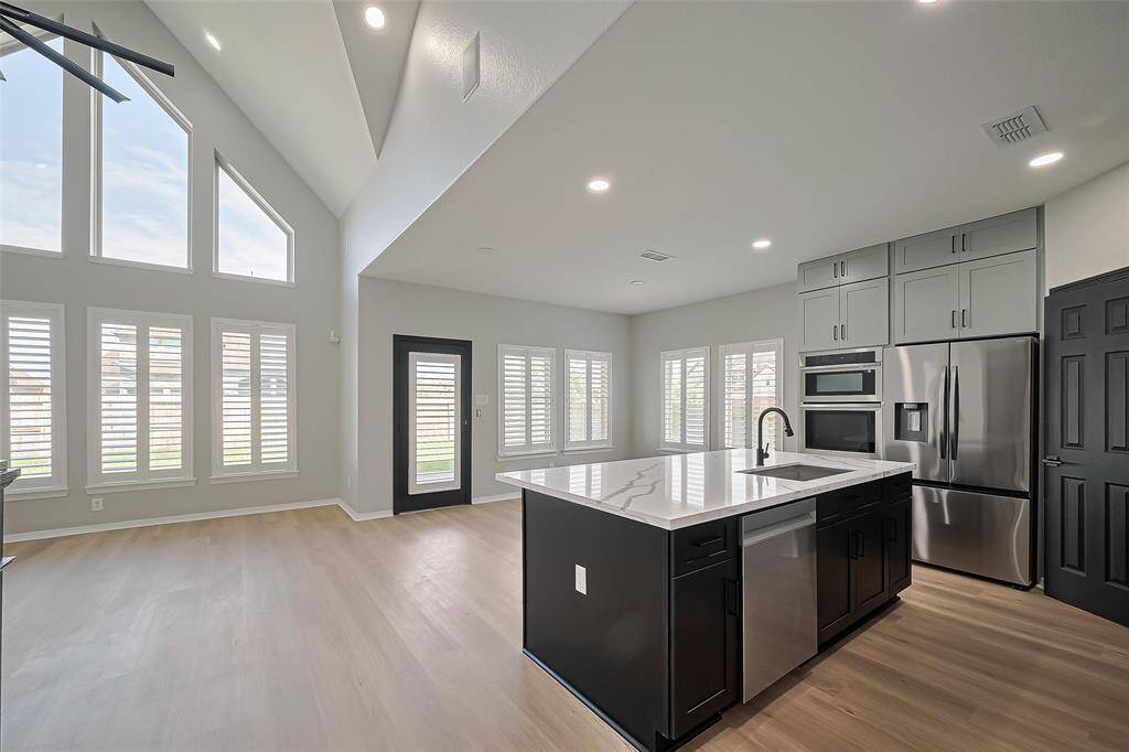 This modern kitchen features an open floor plan with high ceilings and abundant natural light from large windows. It includes a spacious island with a sink, stainless steel appliances, and stylish cabinetry.