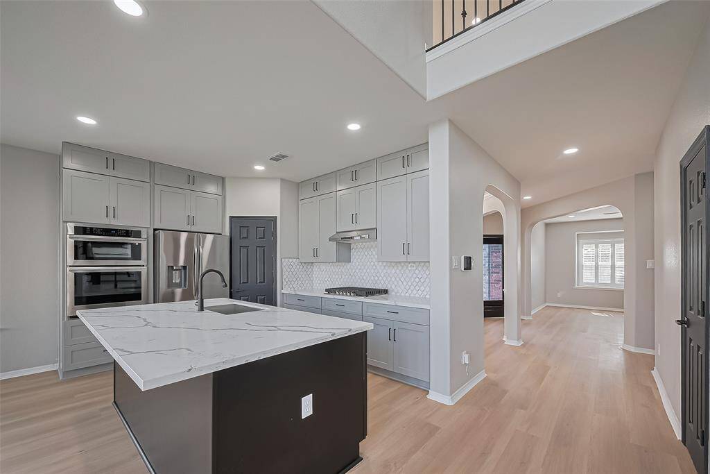 This modern kitchen features sleek gray cabinetry, stainless steel appliances, and a large marble island with a built-in sink. Open layout with bright lighting and an adjacent living space.