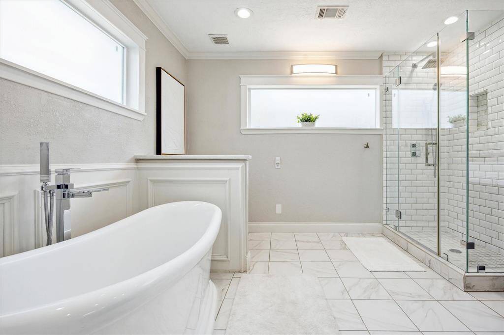 Enjoy the best of both worlds with a luxurious soaking tub and separate glass-enclosed shower, surrounded by elegant wainscoting, marble-look tile, and soft natural light from frosted transom windows.