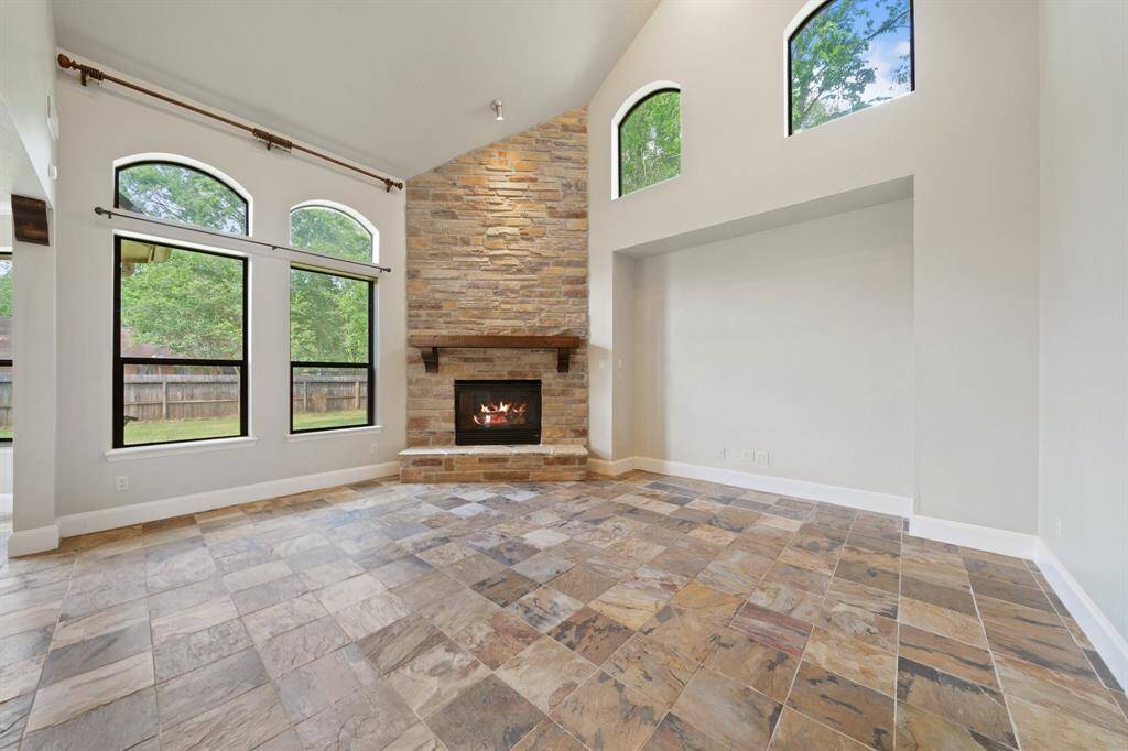 The light brown brick fireplace blends seamlessly with the warm flooring tones, creating a cozy focal point. Two medium windows overlook the yard, filling the room with natural light and a nature view.