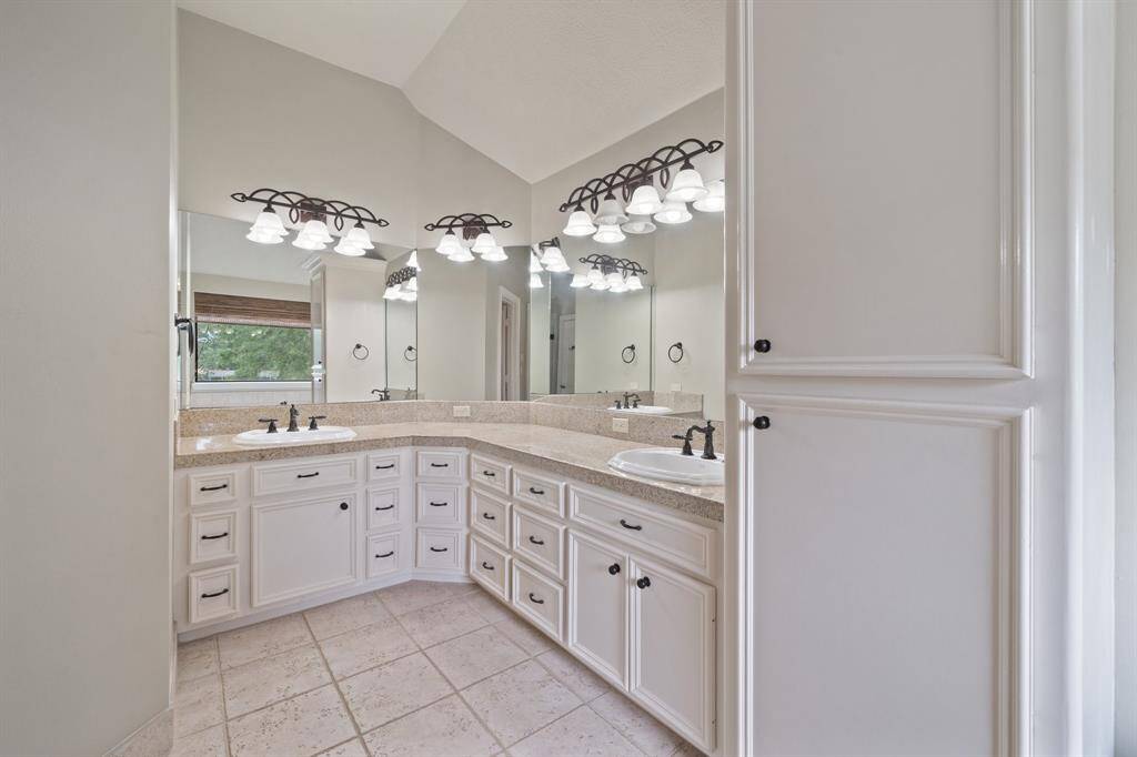 Spacious primary bathroom features crisp white cabinetry, a double corner sink vanity and elegant dark metal light fixtures above the mirrors. The lamp adds and extra charm to this serene, stylish bathroom.