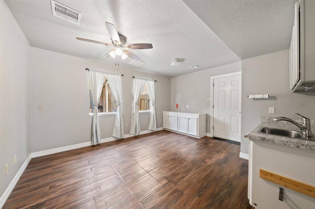 This cozy room, belonging to the lower unit of the garage apartment, is adorned with two large windows with sheer curtains that let in abundant natural light. The space includes a compact kitchenette with granite countertops and white cabinetry, perfect for flexible living.
