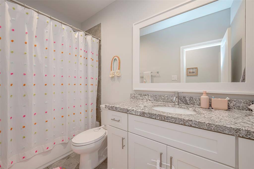 This is a modern bathroom featuring porcelain tile, granite countertop with an under-mount sink, white cabinetry, a large framed mirror, with Shower in Tub. The space has a clean, neutral color palette.