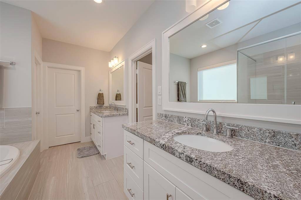 This is a modern bathroom featuring a large vanity with dual sinks and ample storage, sleek granite countertops, and a full-width mirror with overhead lighting. There's a freestanding bathtub to the left and tiled flooring throughout. The space has a neutral sleek look, connected to a walk-in closet, adding convenience and luxury.