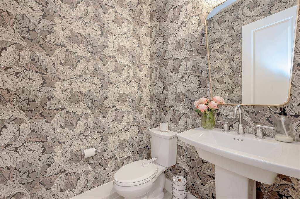 This is a spacious 1/2 bathroom on 1st floor featuring an elegant designer wall paper design, a pedestal sink, a toilet, with a stunning designer mirror.