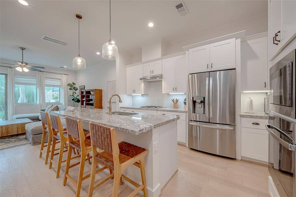 This is a modern, open-concept kitchen featuring white cabinetry, granite countertops, stainless steel appliances, and a breakfast bar. Hardwood floors run throughout, and the space is well-lit with natural and stylish pendant