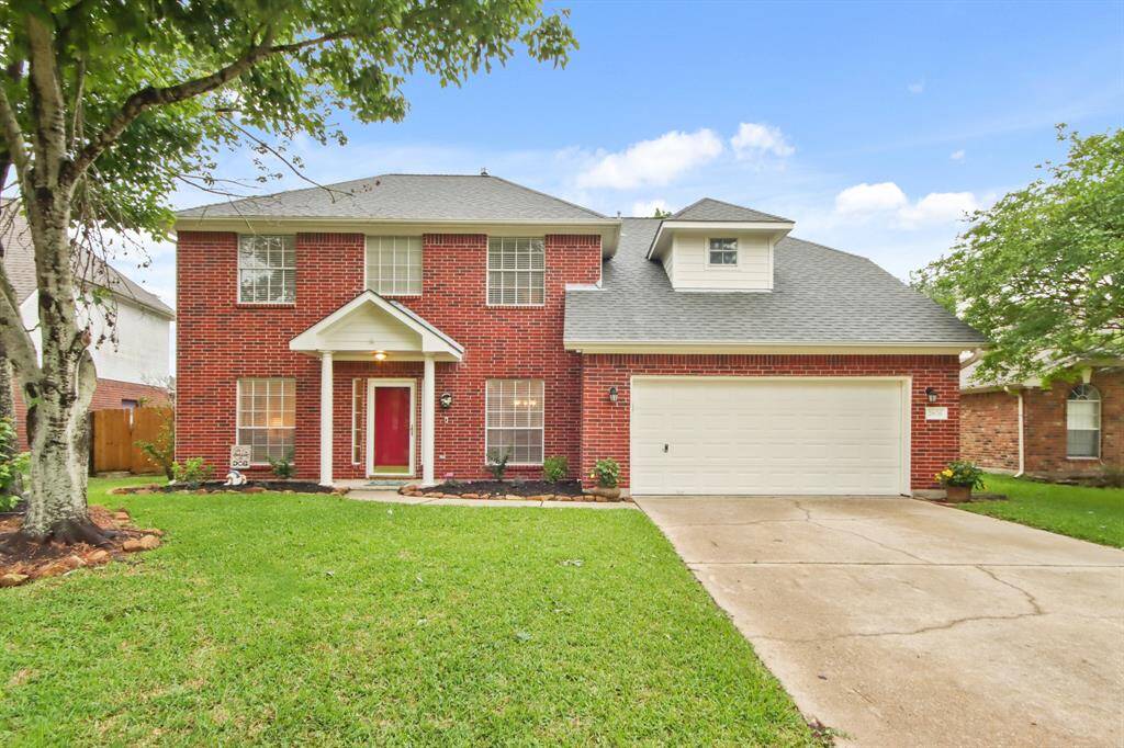 This is a charming two-story brick home featuring a spacious two-car garage, a well-maintained front lawn, and a welcoming red front door. Lush trees and plants enhance its curb appeal.