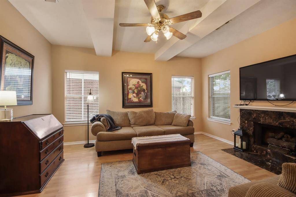 This cozy living room features a warm color palette, a fireplace, large windows with blinds for natural light, wood vinyl flooring,  and a ceiling fan.