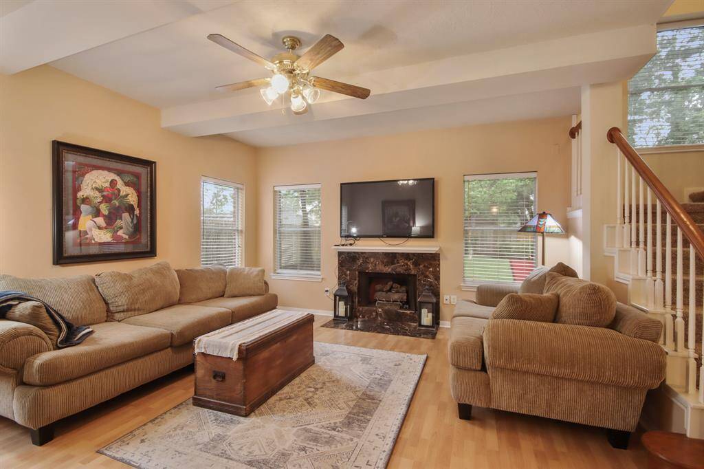 Downstairs cozy living room features wood vinyl plank flooring, with a ceiling fan and fireplace. Large windows offer natural light, and a staircase adds architectural interest.