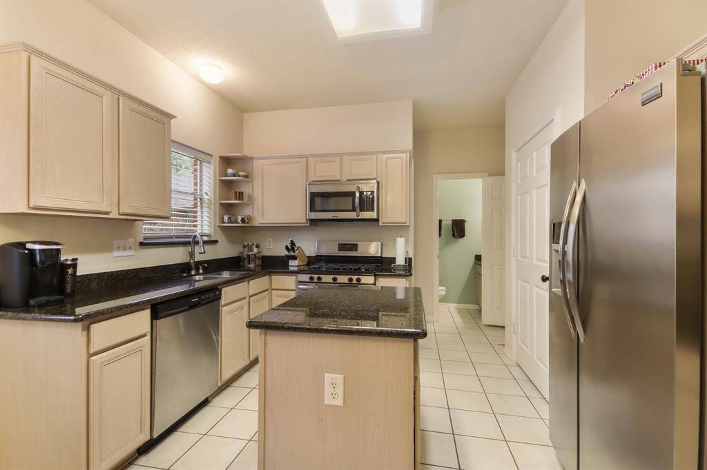This kitchen features light wood cabinetry, stainless steel appliances, island for extra cooking/prepping space. The room is spacious with tile flooring and ample natural light from the window above the sink that looks out into the backyard.