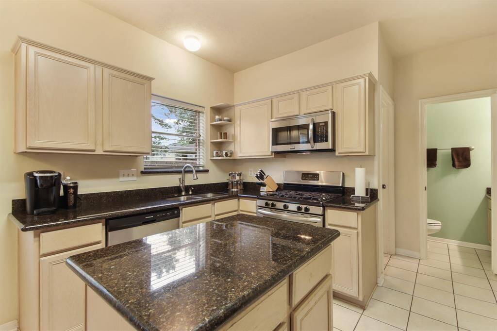 This kitchen features light cabinetry, dark countertops, and stainless steel appliances, including a gas stove and microwave. It has a central island, tiled flooring, and a window for natural light.