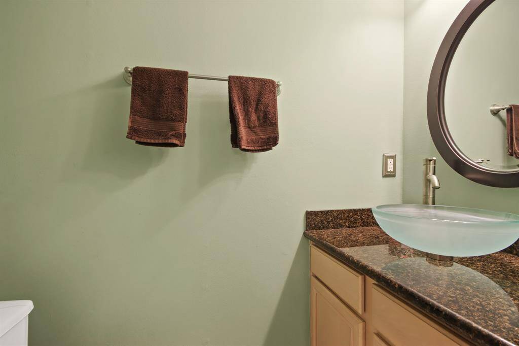 This half bathroom features a modern glass vessel sink on a granite countertop, light green walls and a round mirror, creating a sleek and contemporary space. Located pasted the kitchen next to the laundry area.